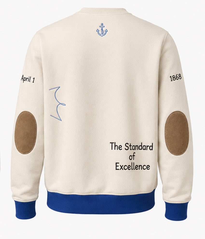 Back view of a cream-colored crewneck for Hampton University. A sweatshirt with blue ribbed cuffs, hem, and collar, featuring brown elbow patches, a blue anchor graphic, and text including 'The Standard of Excellence' and '1868'.