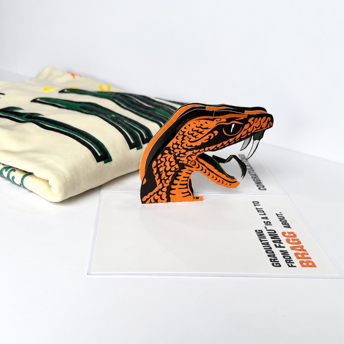 A 3D pop-up of the FAMU mascot, venom, with a folded cream FAMU sweatshirt featuring green and black stripes in the background.