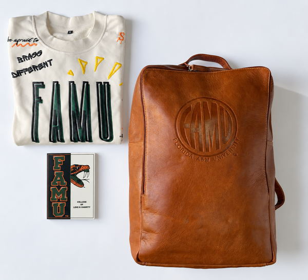 A styled flat-lay on a white background featuring a large buffal leather FAMU backpack, a folded cream FAMU sweatshirt with black and yellow graphics in the upper right, and a small FAMU pop-up card in the upper left.
