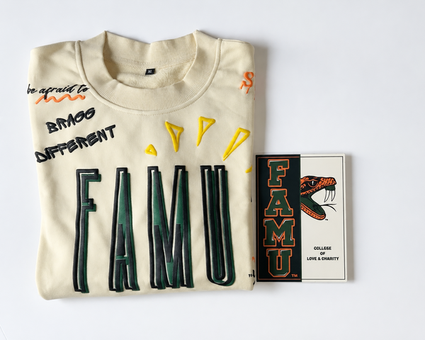 A folded cream FAMU sweatshirt with bold black, green, and yellow graphic text and shapes, displayed on a white background next to a small printed FAMU card featuring the same graphic design.