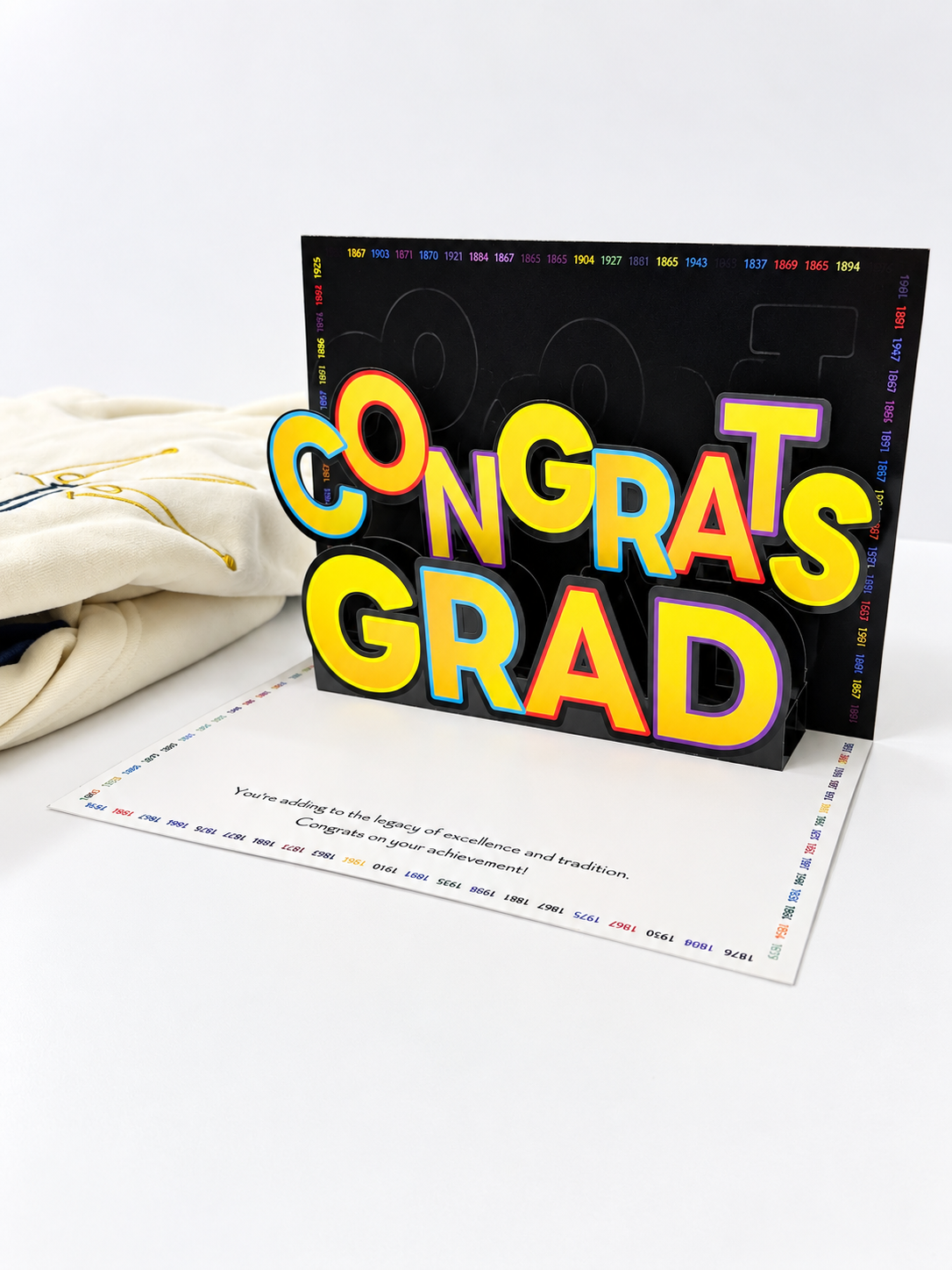 A pop-up congratulations card for an HBCU student, with a folded cream fabric HBCU hoodie beside it. The card features large 3D text reading 'CONGRATS GRAD' in yellow, orange, and blue with a black background.