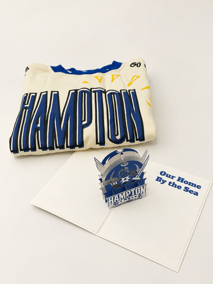 A folded cream-colored t-shirt with bold blue and navy 'HAMPTON' lettering and yellow accents, displayed next to a 3D pop-up card featuring a matching blue and silver HAMPTON ship graphic and the text 'Our Home By the Sea'.