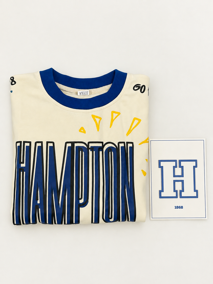 A folded cream-colored Hampton 1968 t-shirt with a blue collar and large blue 'HAMPTON' text outlined in black, displayed next to a small white card featuring a blue 'H' and '1968'.