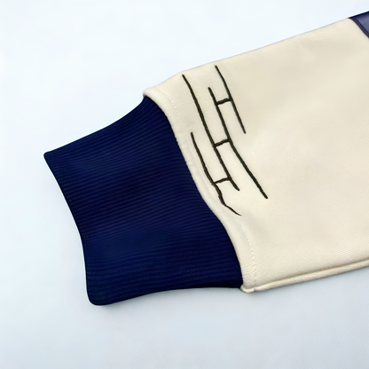 A close-up of a ribbed knit sleeve or cuff in cream and navy blue, featuring a black embroidered geometric logo on the cream section for the HBCU Legacy hoodie.